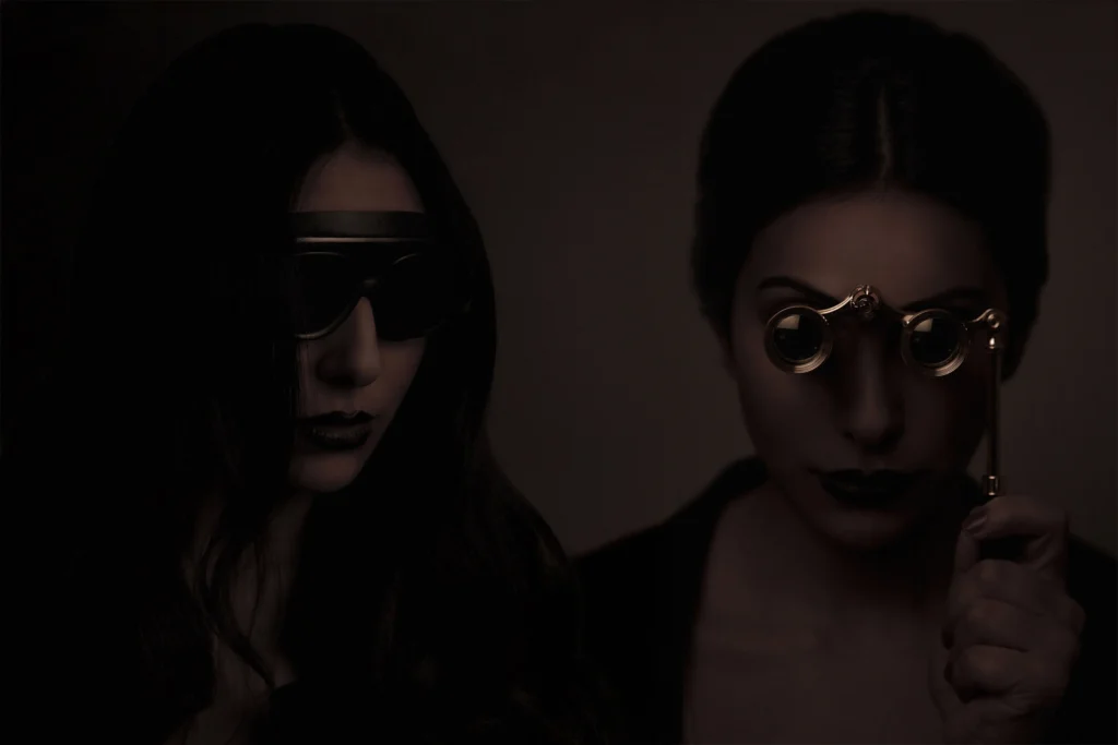 Portrait éditorial en studio de deux femmes avec masque et jumelles, ambiance sombre inspirée d’un univers occulte, série Coven créée par Armando Garcia