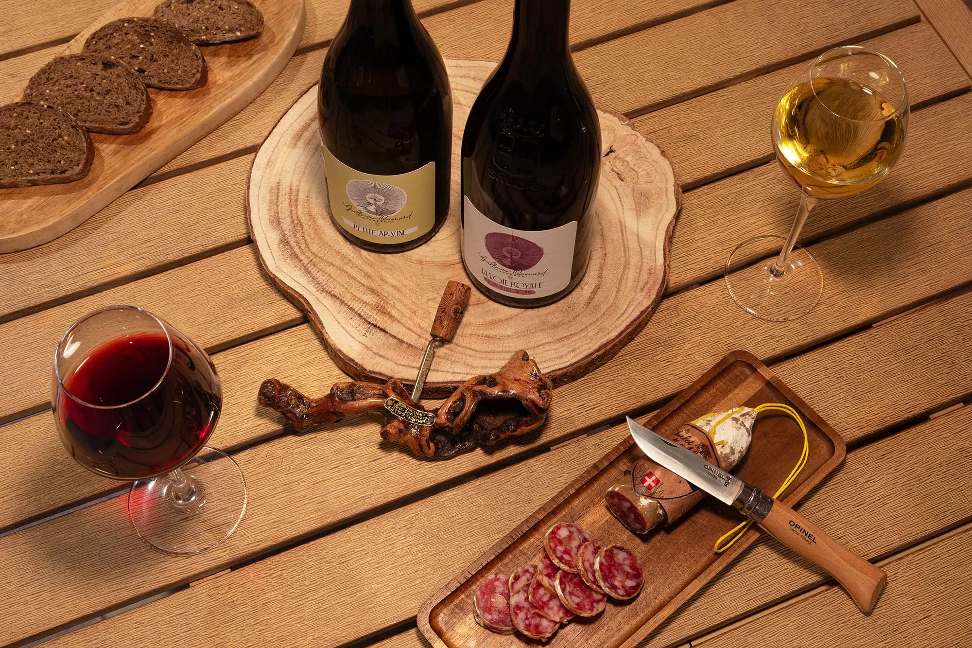 Photographie d'art de vivre, nature morte aux vins de Savoie, bouteilles, verres, saucisson et couteau Opinel sur table en bois, Hôtel Le Refuge Urbain Kyriad à Chambéry, par Armando Garcia